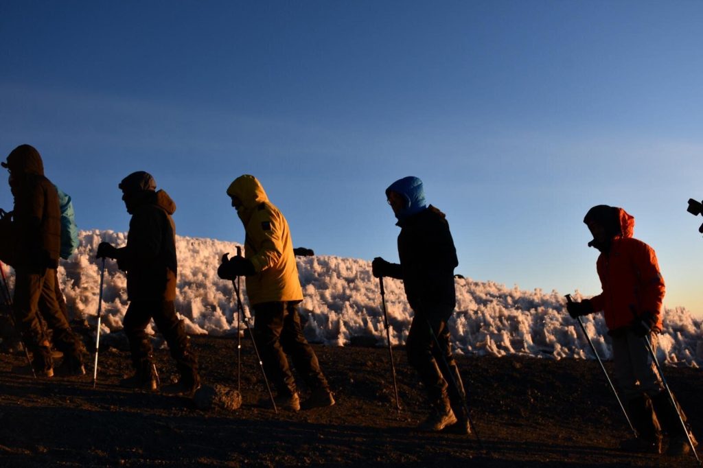 Five trekkers silhouetted beside a dawn-lit ice wall on the crater rim of Kilimanjaro