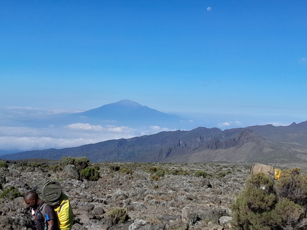Porter carrying a load across open moorland with a distant volcanic cone behind on Mount Kilimanjaro