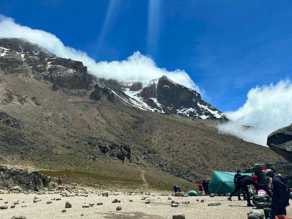 Porters and a green mess tent at a camp below snow streaked cliffs on Mount Kilimanjaro