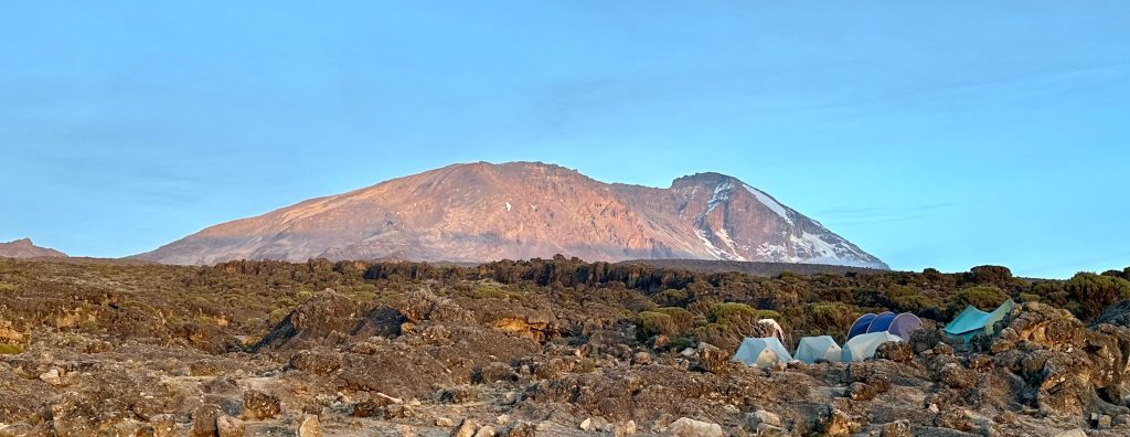 Tents pitched on open ground below a sunlit snow streaked summit dome on Mount Kilimanjaro