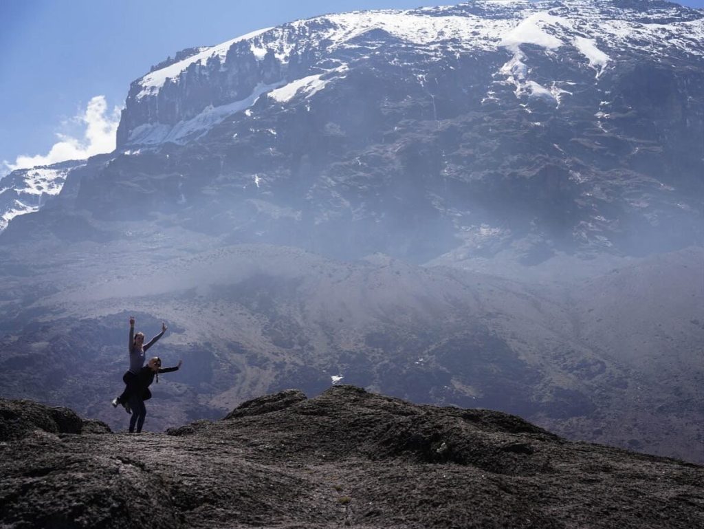 Two women cheering on a rock below the snowy cliff on Mount Kilimanjaro